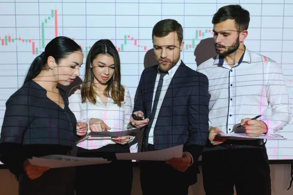 Four business professionals stand together reviewing financial statements, with a financial chart projected on the wall behind them. They appear focused, discussing finance and accounting teams' strategies and how to improve month end closing process.