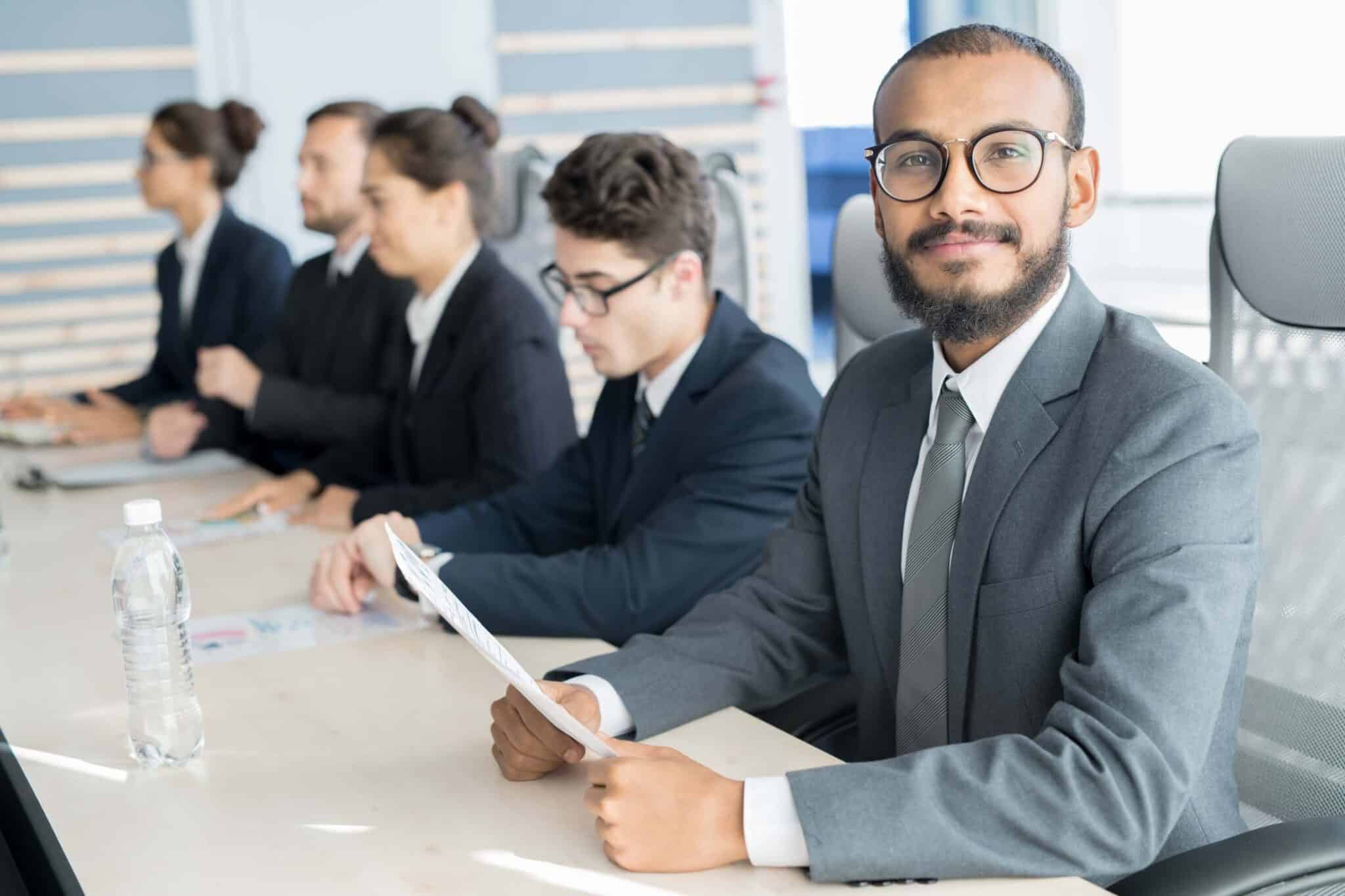 A young man in a suit and glasses sits at a conference table holding papers, smiling at the camera. Three colleagues in business attire are beside him, discussing how to improve month end closing process as they review their documents.