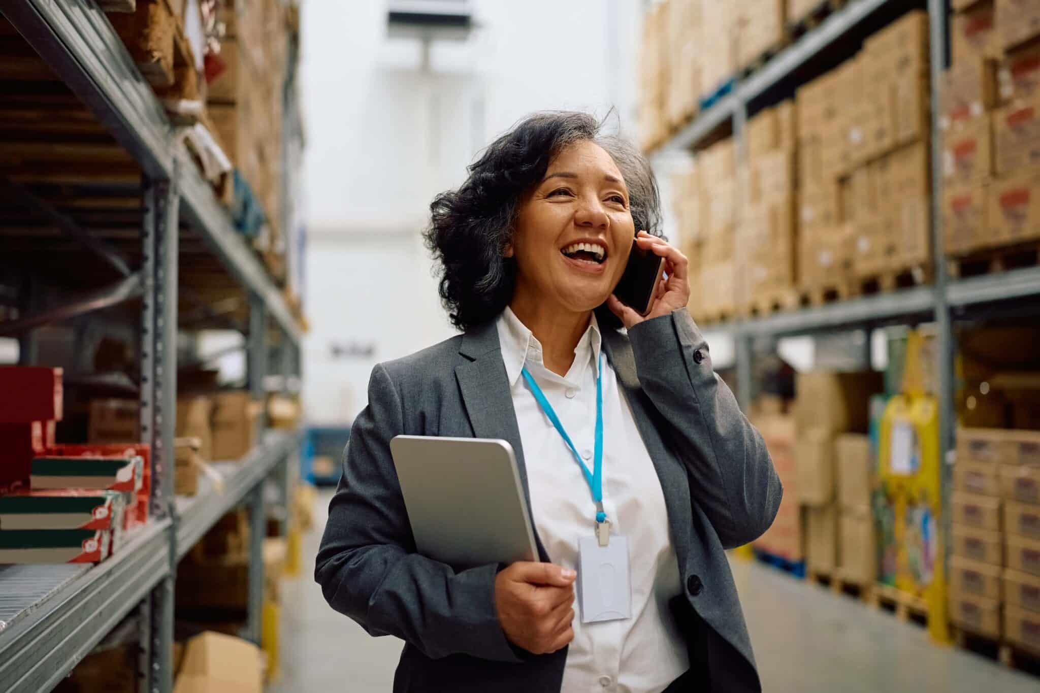 A smiling woman in a suit holding a tablet and talking on her phone stands between shelves in a warehouse filled with boxes, efficiently managing inventory with wholesale accounting software.