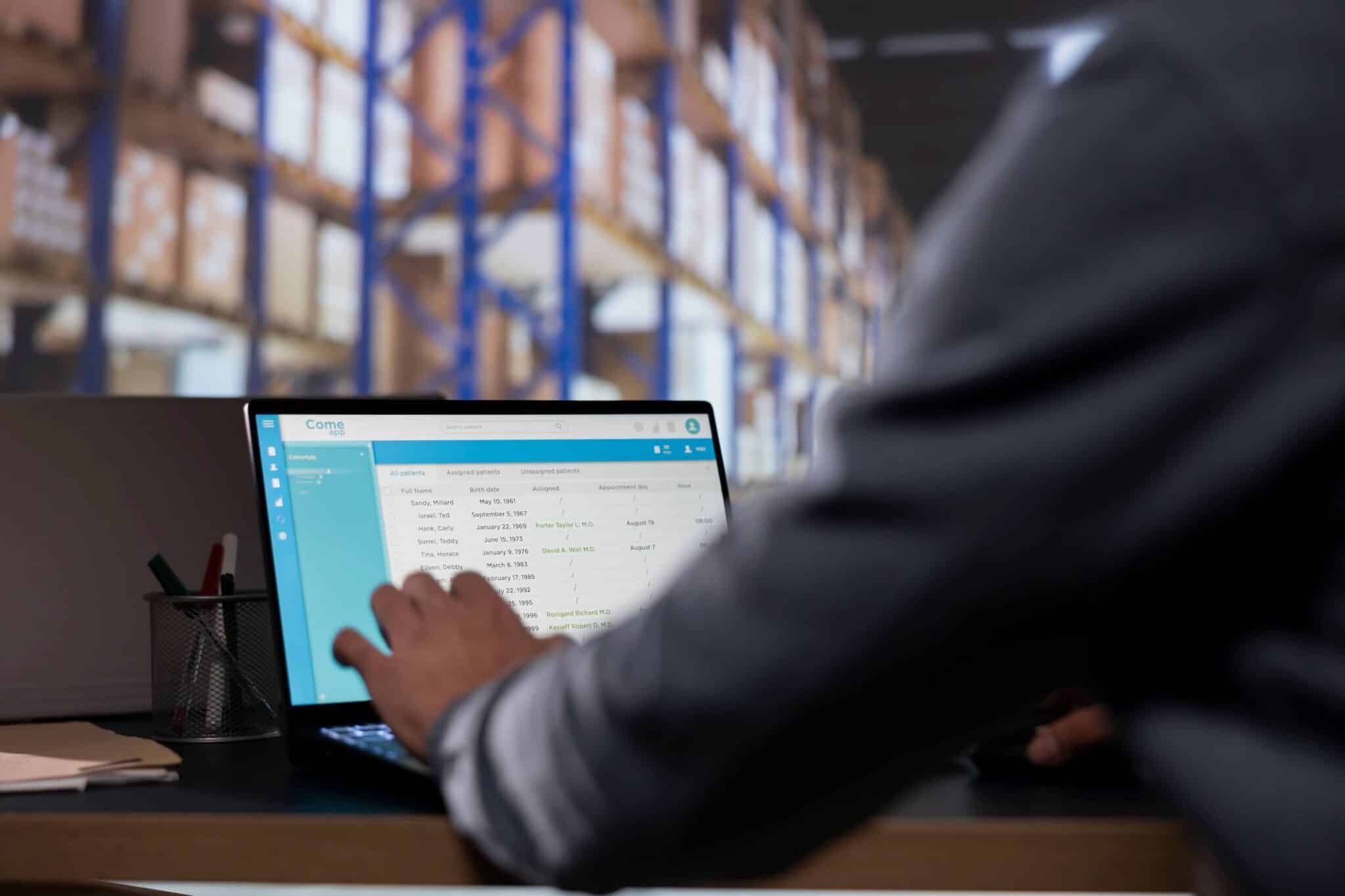A person using a laptop at a desk with wholesale accounting software and an inventory management system displayed on the screen; blurred shelves and boxes in a warehouse are visible in the background.