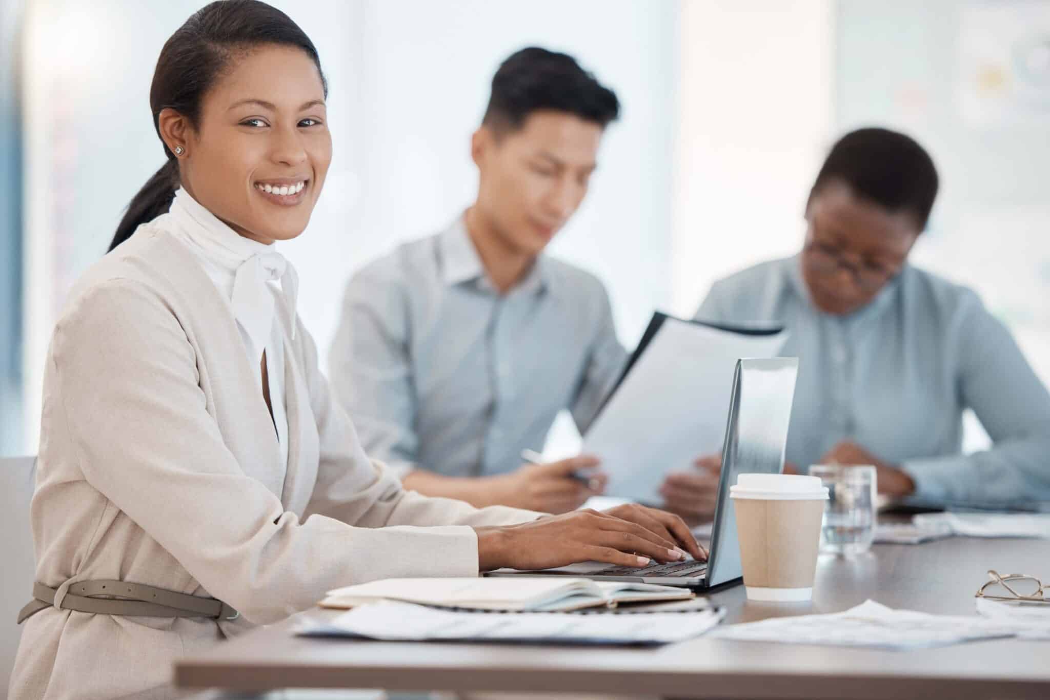 A woman smiles at the camera while working on a laptop with Deltek Replicon payroll time tracking software, as two colleagues in the background focus on documents. A coffee cup and papers are on the table.