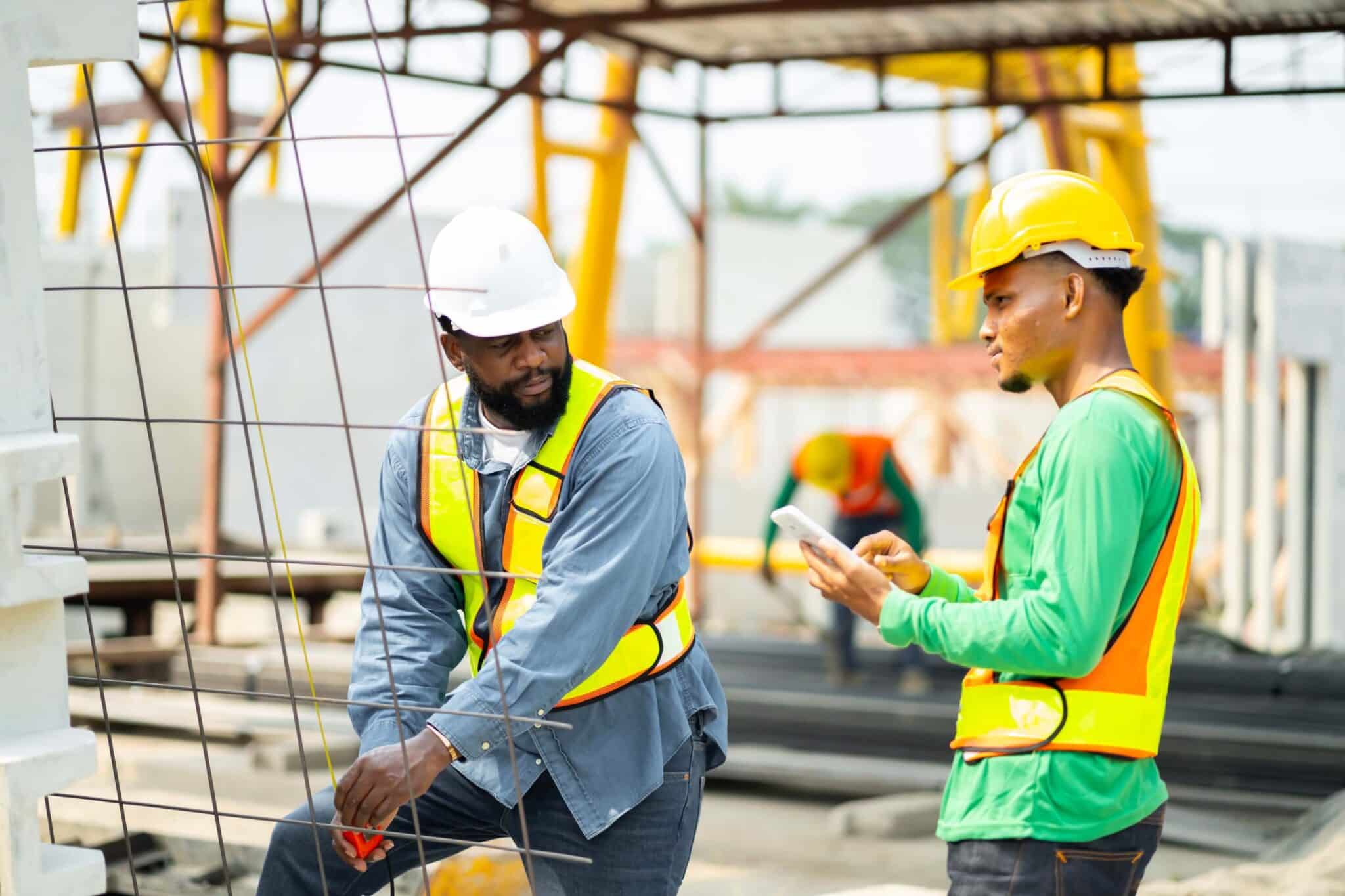Two construction workers wearing safety helmets and reflective vests discuss plans at a building site, using a tablet with Replicon mobile time tracking software. Construction materials and another worker are visible in the background.