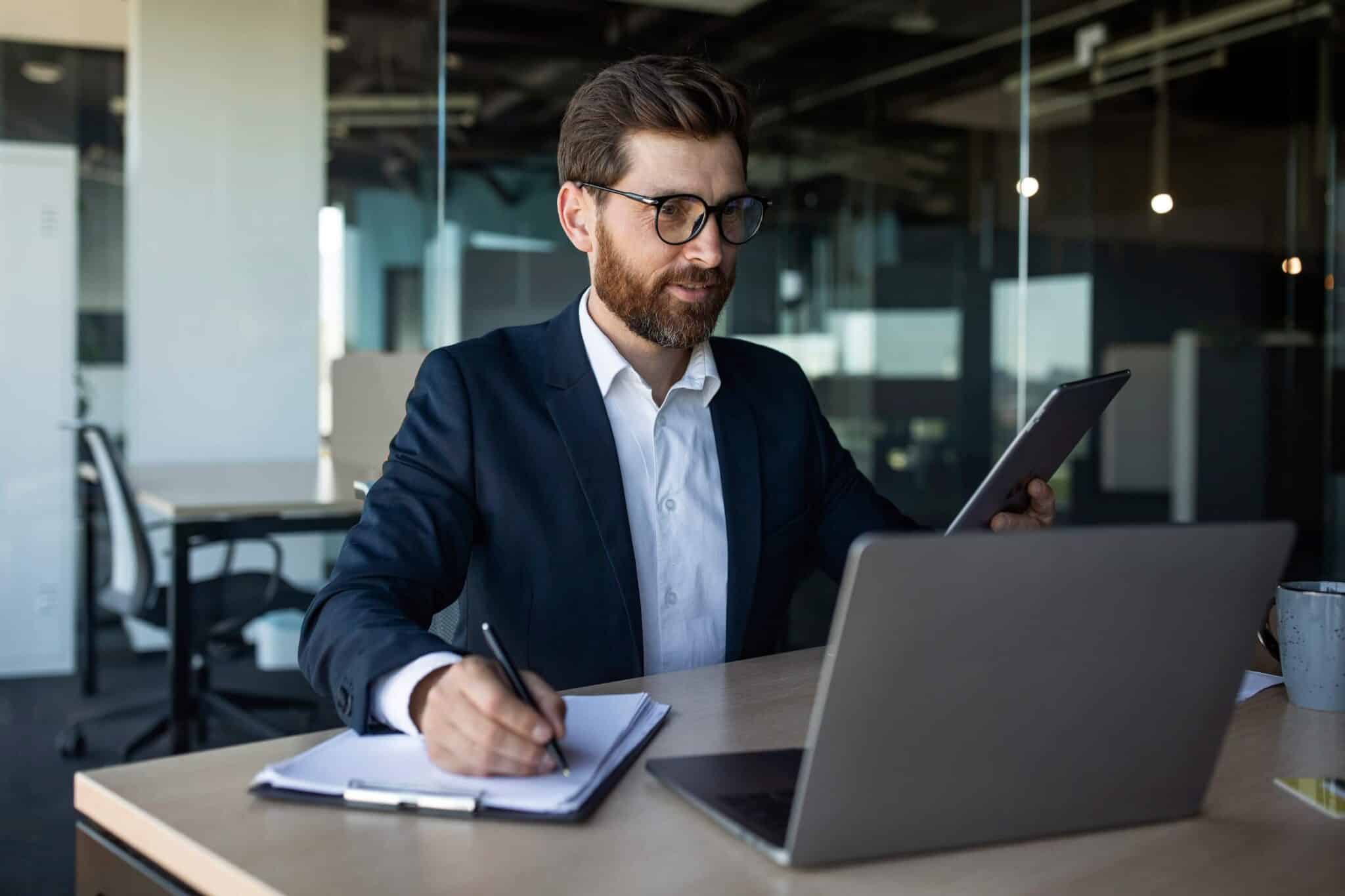 A man in a suit and glasses sits at a desk in a modern office, holding a tablet displaying wholesale accounting software in one hand and writing on a clipboard with the other, with an open laptop and a mug nearby.