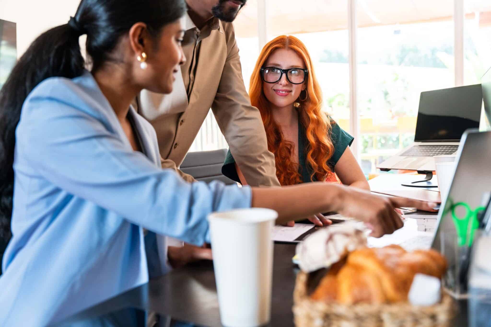 Three people in business attire discuss at a desk with laptops, documents, a coffee cup, and pastries. One woman points at the laptop displaying Replicon mobile time tracking software while another looks on and a man stands nearby.