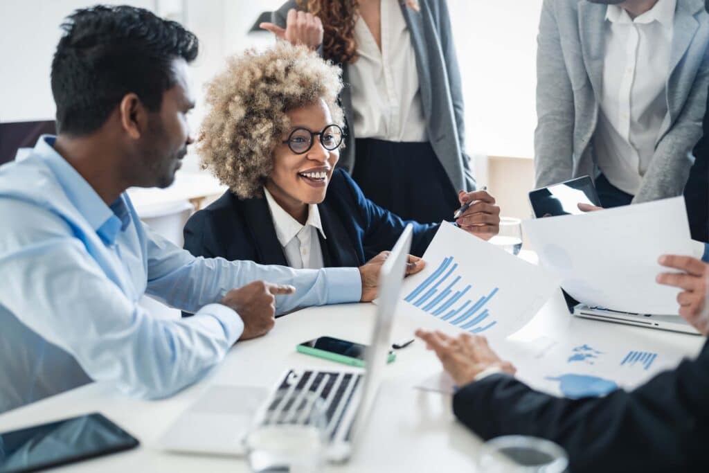 A group of business professionals sits around a table, discussing charts, graphs, and how to improve cash flow forecast. A woman with curly hair and glasses smiles while listening. Laptops, documents, and phones are visible on the table.