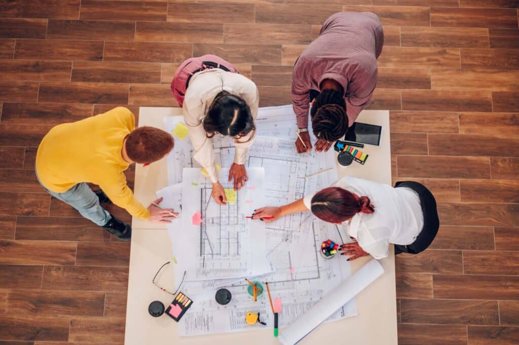 Four people stand around a table covered with architectural blueprints and sticky notes, collaborating and discussing project margins as they point at the plans. Various office supplies and coffee cups are also on the table.