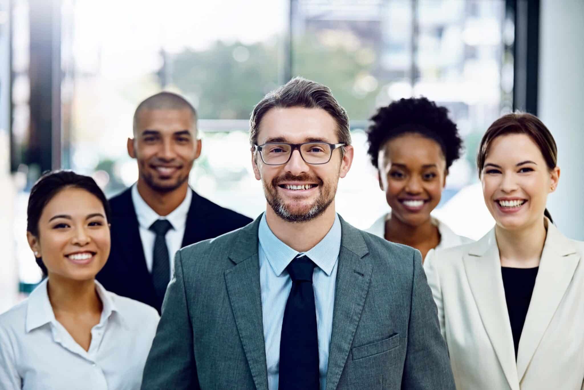 A group of five professionally dressed people, three women and two men, stand close together indoors, smiling confidently at the camera—ready to discuss how to improve cash flow forecast. The background is bright and slightly out of focus.