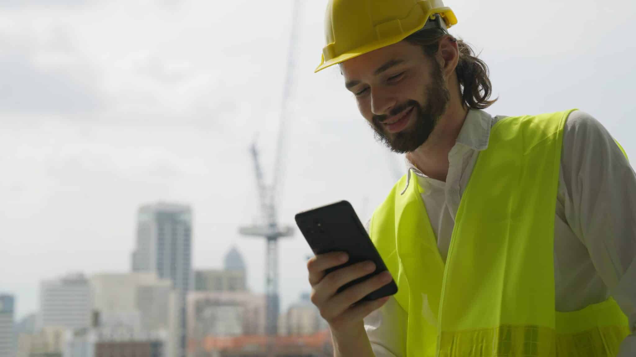 A smiling construction worker in a yellow safety vest and hard hat checks his smartphone, using Replicon mobile time tracking software. City buildings and a crane are visible in the blurred background.