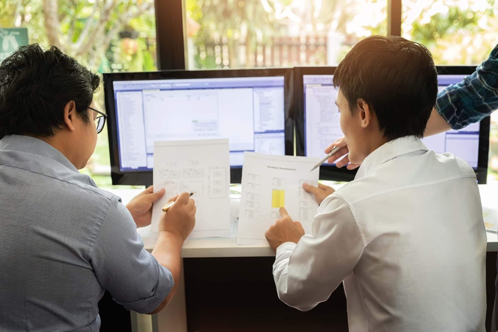 Two people sit at a desk, reviewing flowcharts and documents while discussing Replicon mobile time tracking software. Three monitors show diagrams and code in the background as a third person, partially visible, points at the papers.