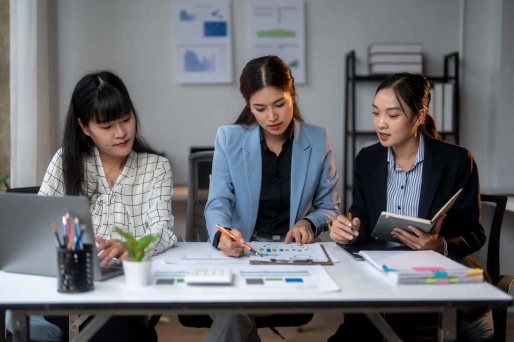 Three women in business attire sit at a desk with charts, documents, and a laptop discussing budget variance analysis. One writes on a clipboard, another types on a laptop, and the third holds a notebook.