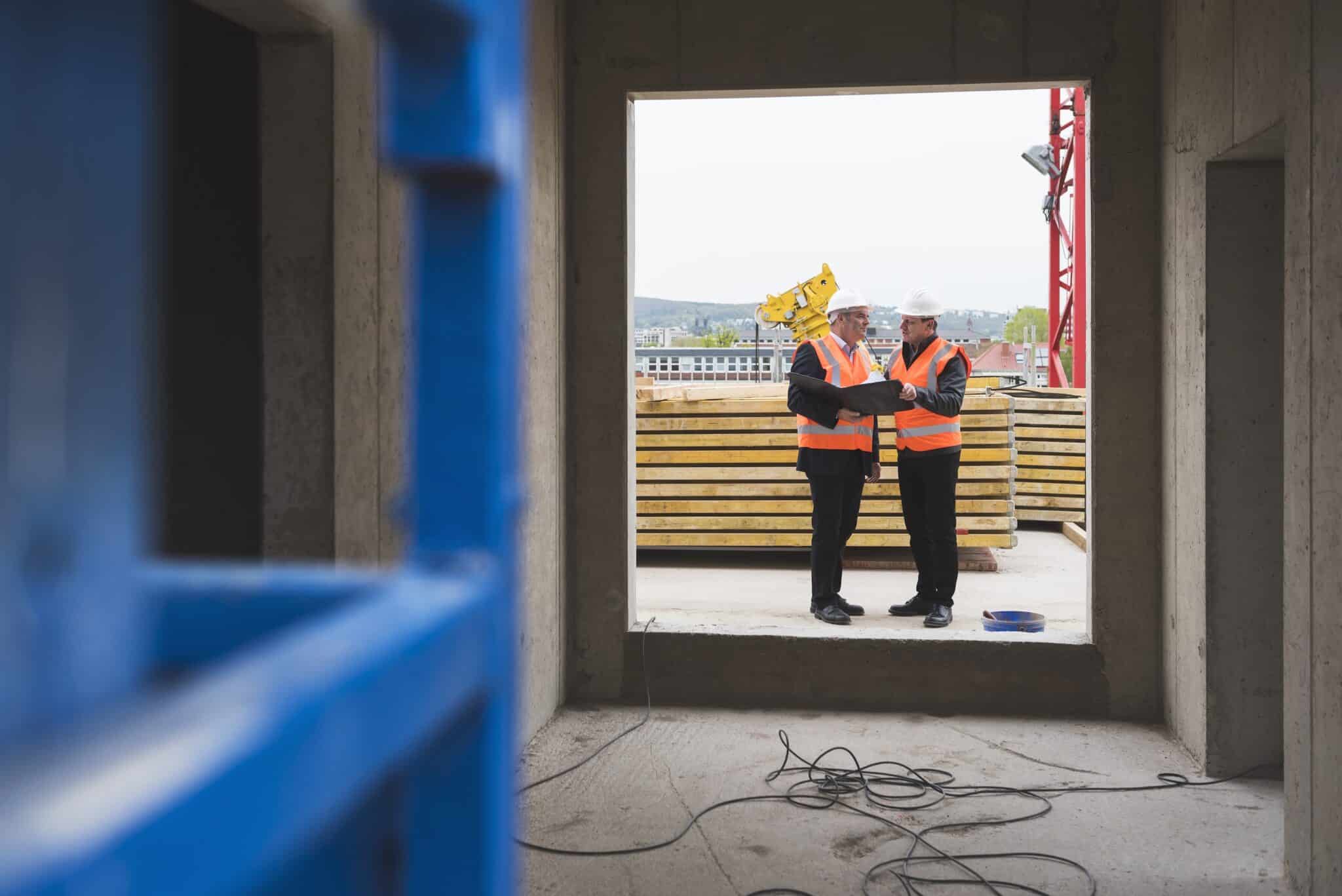 Two construction workers in orange safety vests and helmets stand outside a building frame, reviewing blueprints. Wooden boards, a crane, and loose cables are visible—highlighting the need for Deltek Replicon payroll time tracking software on site.