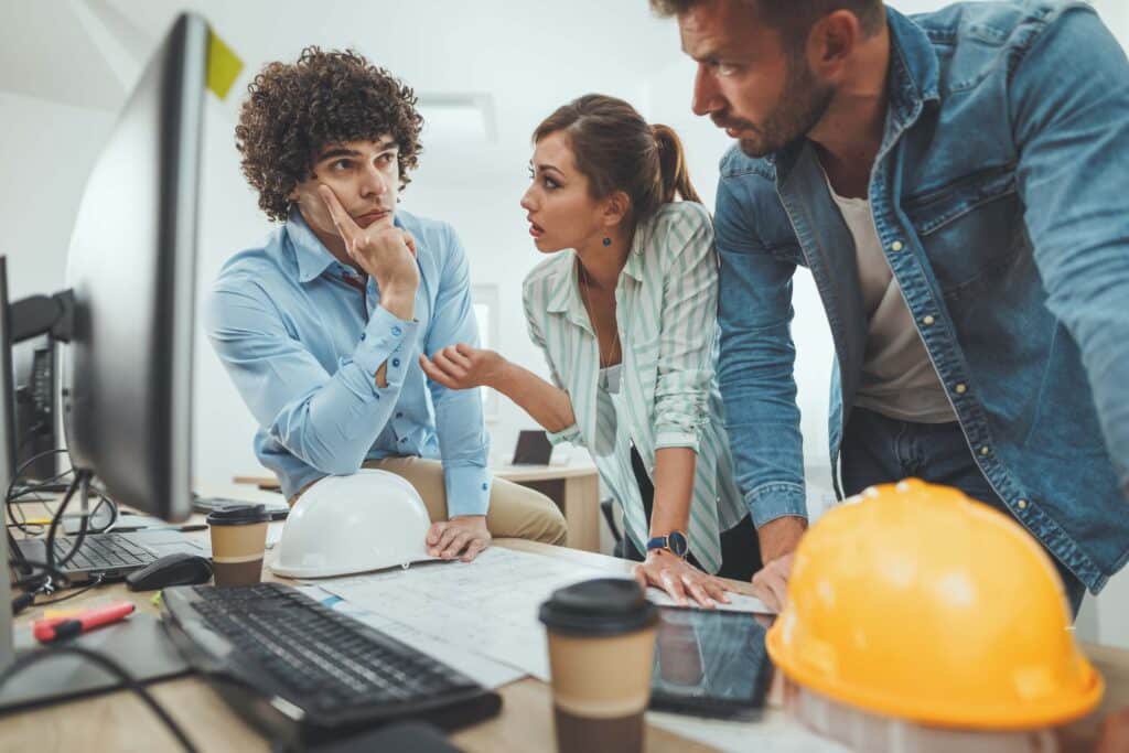 Three colleagues discuss project management at a desk with blueprints, a computer, and safety helmets. One man looks thoughtful—perhaps pondering why most projects are still late despite contingency planning—as the others talk intently.