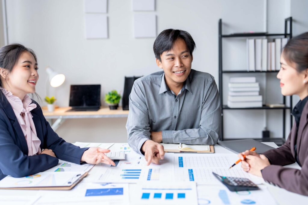 Three people in business attire sit at a table covered with charts, graphs, and documents discussing budget vs actual variance in a modern office. One man smiles while speaking, and the two women listen attentively.