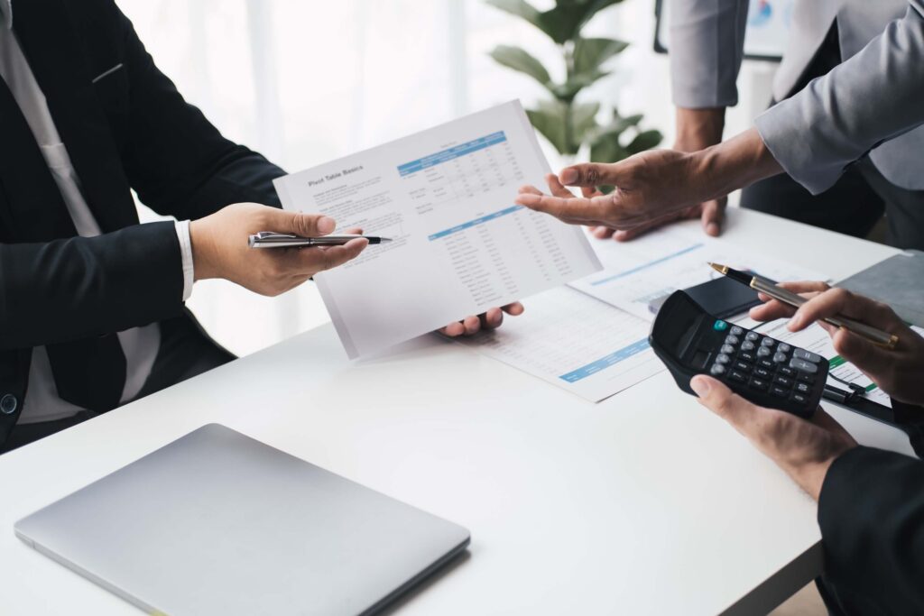 Three people at a desk hold documents, a calculator, and a pen while discussing financial statements. A closed laptop sits beside them as finance and accounting teams collaborate, with plants visible in the background.