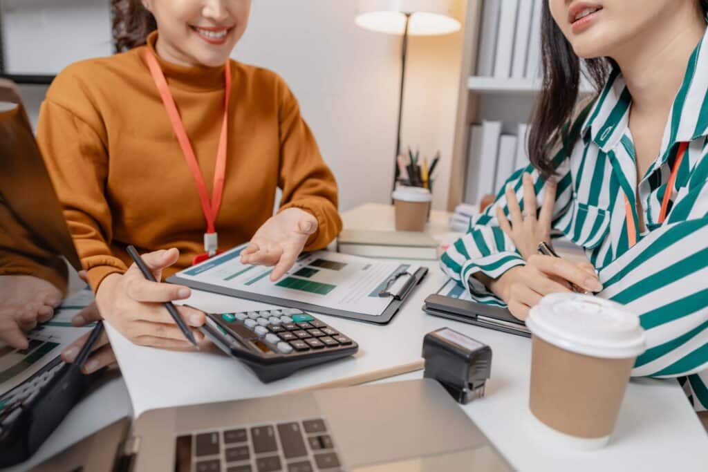 Two women sit at a desk discussing documents with charts related to intercompany transactions, one gesturing while holding a clipboard and a calculator. Coffee cups, a laptop, and office supplies are visible on the desk.