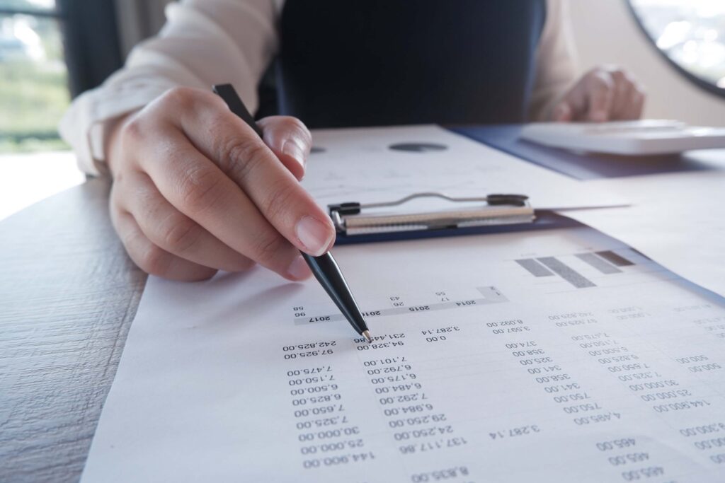 A person holds a pen and points at numbers on a financial document with charts, while other papers and a clipboard are visible on the desk, possibly reviewing details during the invoice matching process.