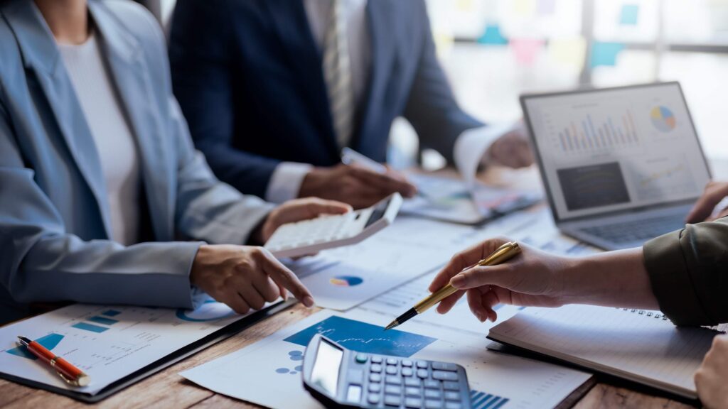 Three people in business attire discussing intercompany transactions at a table, using calculators, a laptop, and reviewing charts and graphs with pens and notepads.