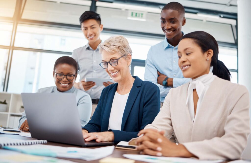 A diverse group of five professionals, three women and two men, gathered around a laptop in a bright office, smiling and collaborating on consolidated financial statements for their project.