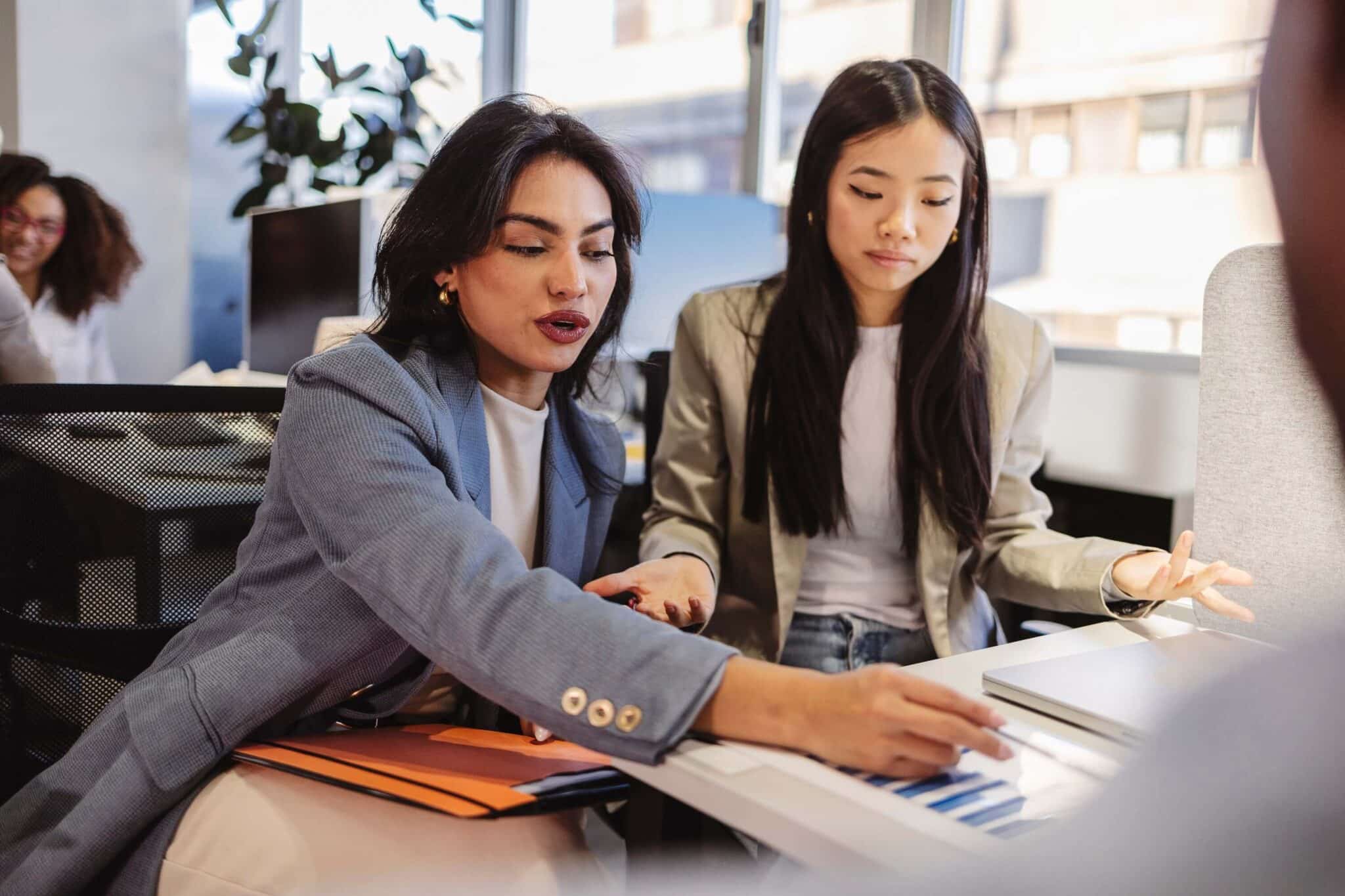 Two women in business attire sit at a desk in an office, reviewing documents on how to eliminate intercompany transactions in consolidation. One gestures toward the papers while the other listens attentively, with bright windows and coworkers in the background.