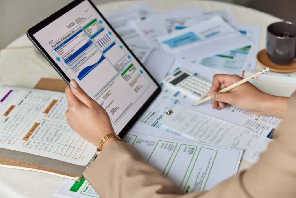 A person reviews data on a tablet and writes notes on paper documents spread across a table, analyzing paperwork, invoices, and a calculator—suggesting a business setting focused on the invoice matching process and purchase order verification.