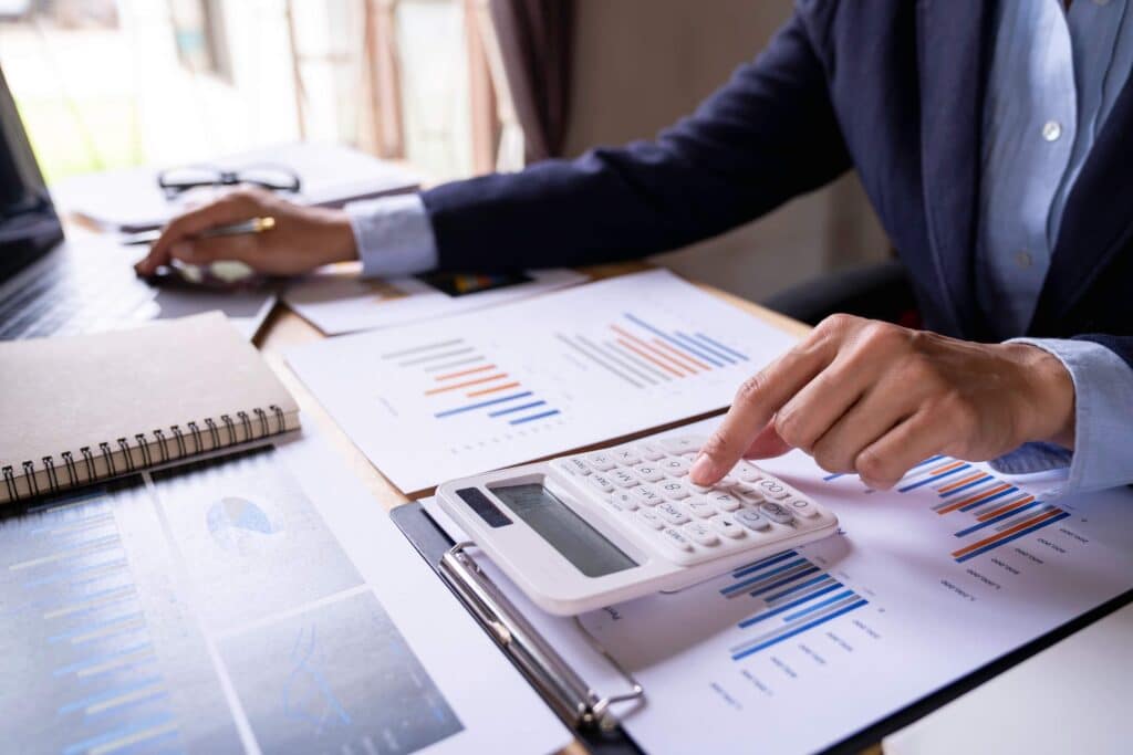 A person in business attire uses a calculator at a desk with financial charts, graphs, a notebook, and a laptop, analyzing intercompany transactions and making calculations for multiple legal entities.