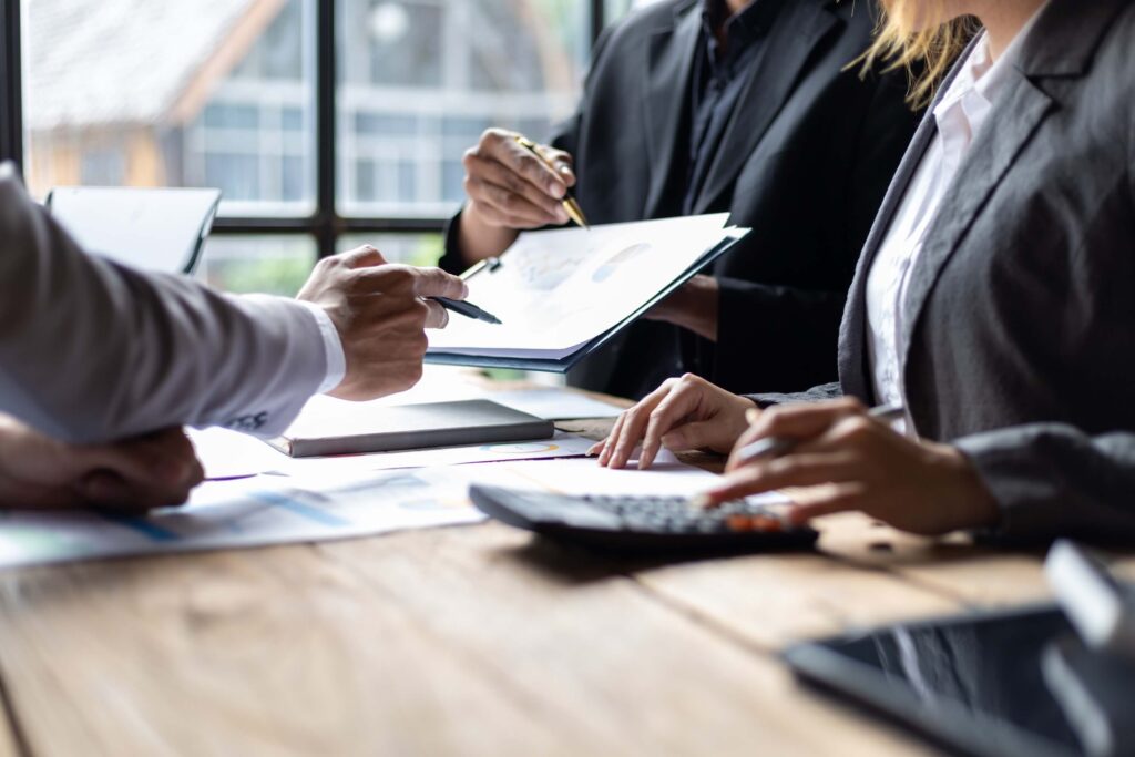Three people in business suits sit at a desk with documents, a calculator, and a tablet, discussing consolidated financial statements and intercompany eliminations in a well-lit office setting.
