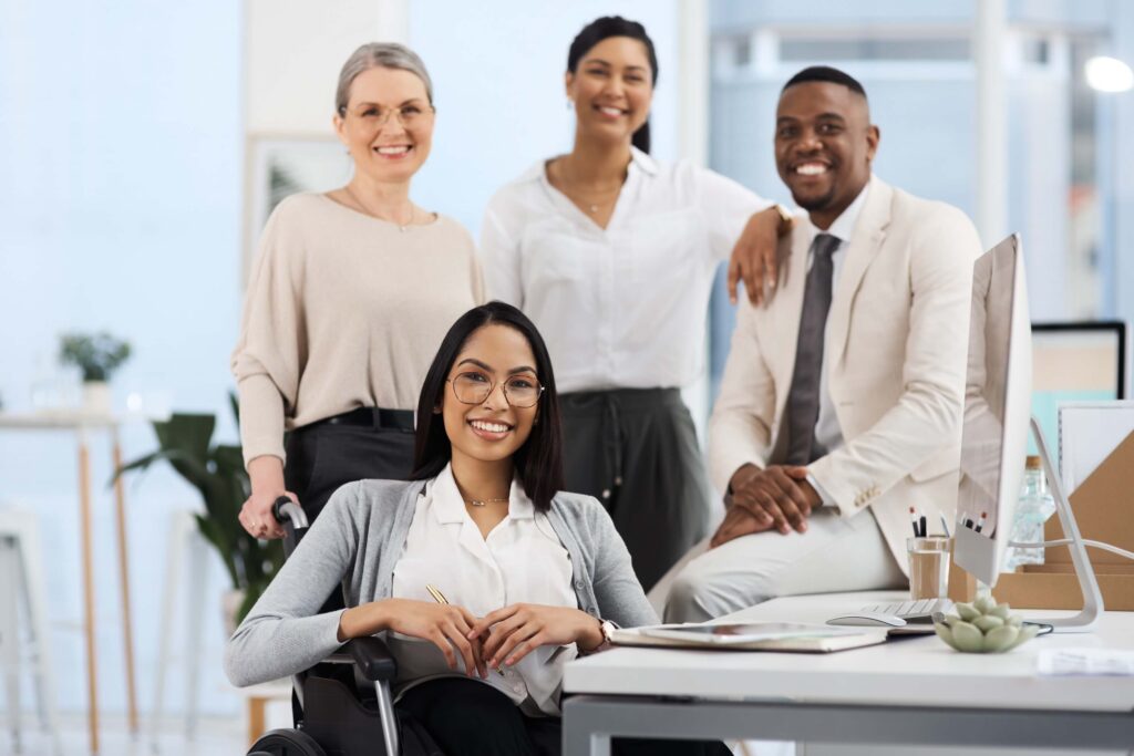 Four professionally dressed colleagues, including a woman in a wheelchair, smile together in a modern office setting, conveying diversity and teamwork—values essential when collaborating on consolidated financial statements.