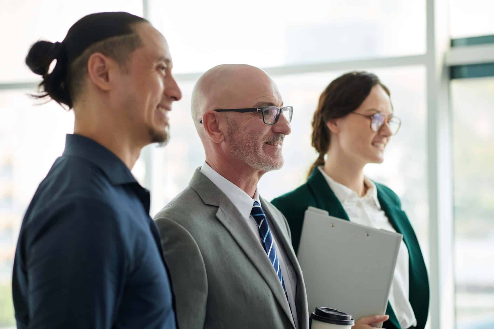 Three business professionals, two men and one woman, stand side by side smiling in a bright office. The woman holds a clipboard and coffee cup, possibly discussing why an invoice does not match purchase order with her colleagues.