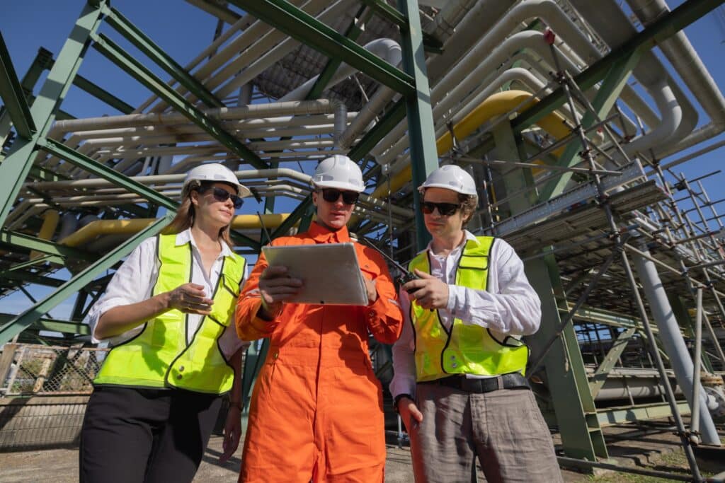 Three engineers in hard hats and safety vests stand outdoors at an industrial plant, reviewing project management details on a tablet together in front of large metal pipes and structural framework.