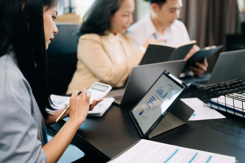 Three people work at a table with laptops, notebooks, and documents. A woman in the foreground uses a calculator and views consolidated financial statements on a tablet, while two others discuss notes in the background.