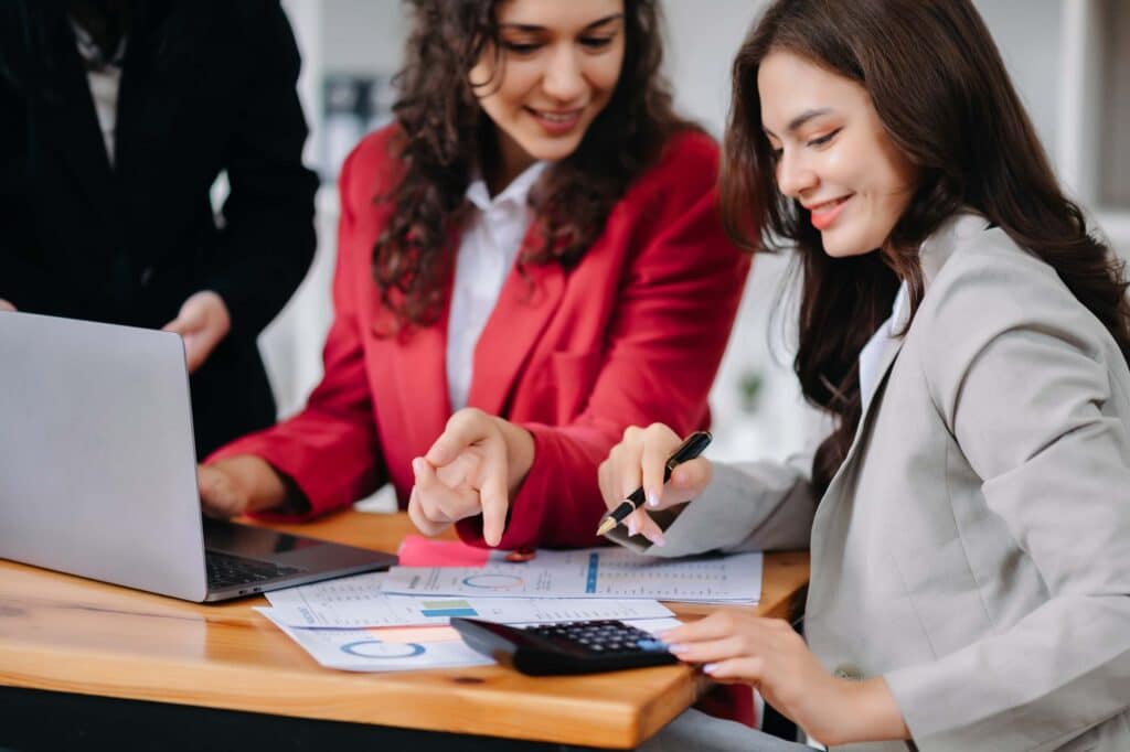 Two women in business attire sit at a desk, smiling as they discuss financial documents with charts and graphs. One uses a calculator while the other points to papers, reviewing the invoice matching process with their laptop open on the table.