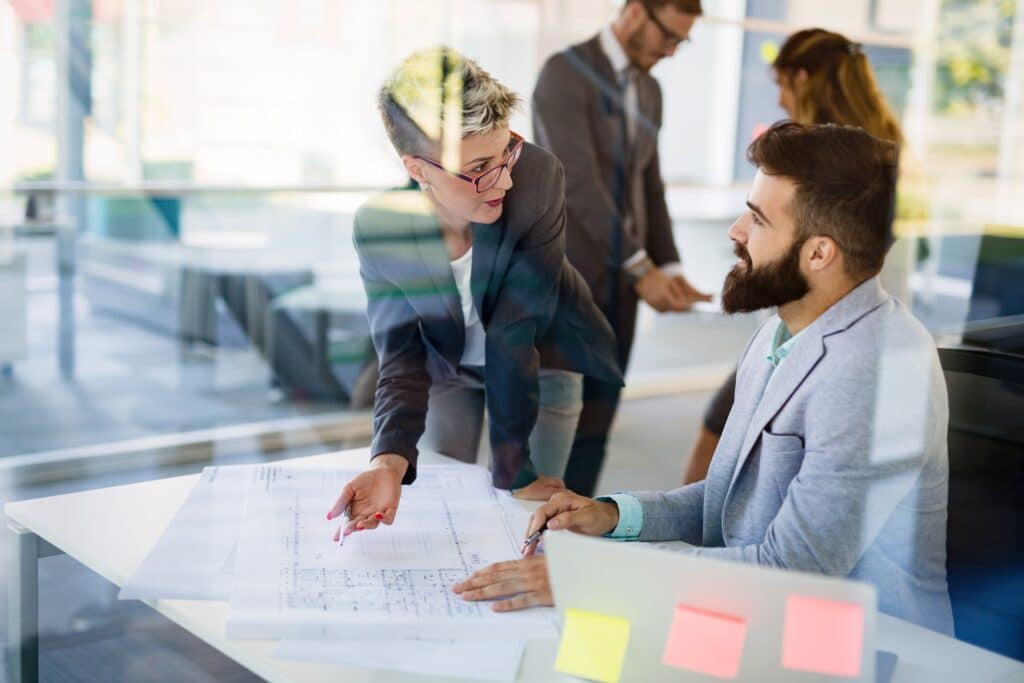 Two colleagues in business attire discuss architectural plans at a desk in a modern office, focusing on project management strategies to manage scope creep. Laptops, documents, and sticky notes help organize their efforts as others work in the background.