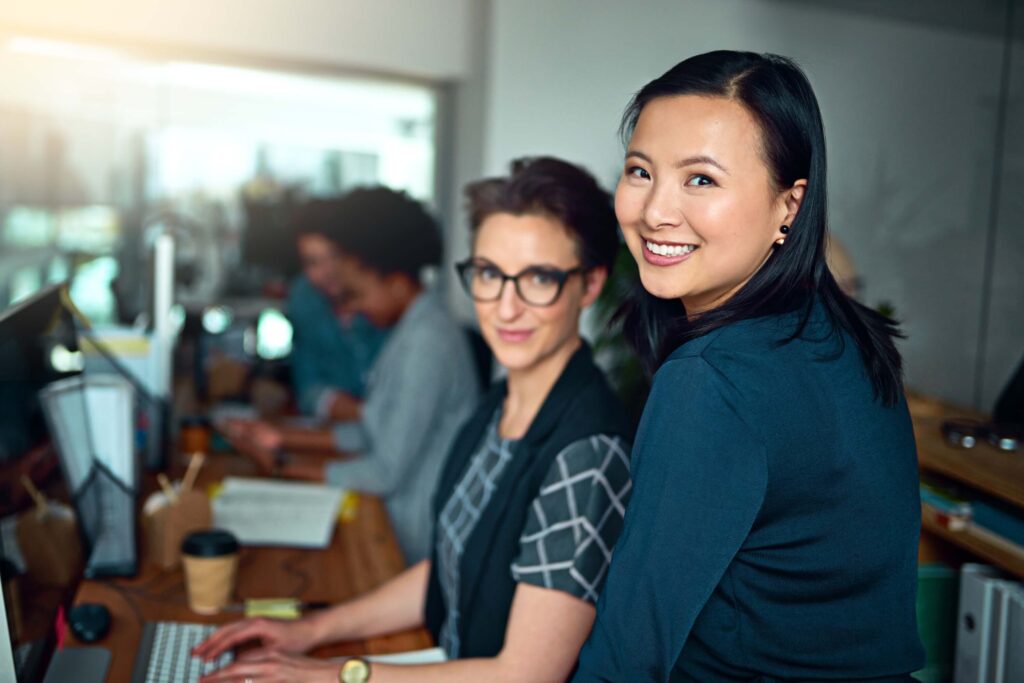 Two women in an office, one typing at a desk and the other standing beside her, smiling at the camera. The teamwork behind them highlights their collaboration on intercompany accounting and managing legal entities.