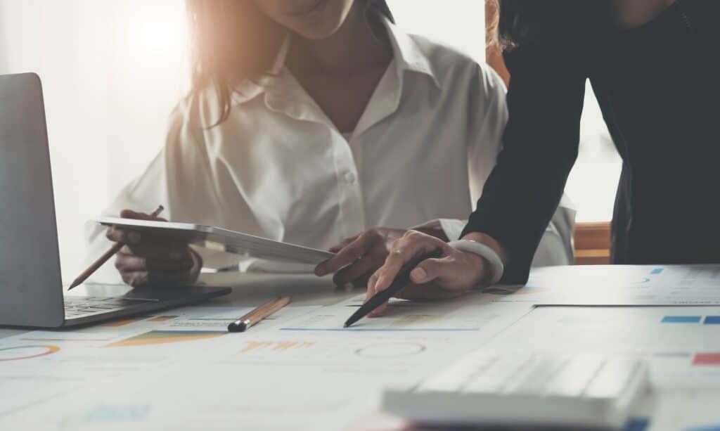 Two people work together at a desk covered with documents and charts; one holds a tablet while the other points at a paper, suggesting collaboration on project management or how to prevent scope creep in business or financial data.