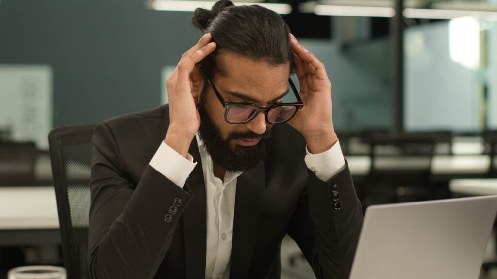 A man in a suit and glasses sits at a desk with a laptop, holding his head with both hands and looking stressed, perhaps overwhelmed by title 31 casino compliance or the demands of financial crimes enforcement network regulations.