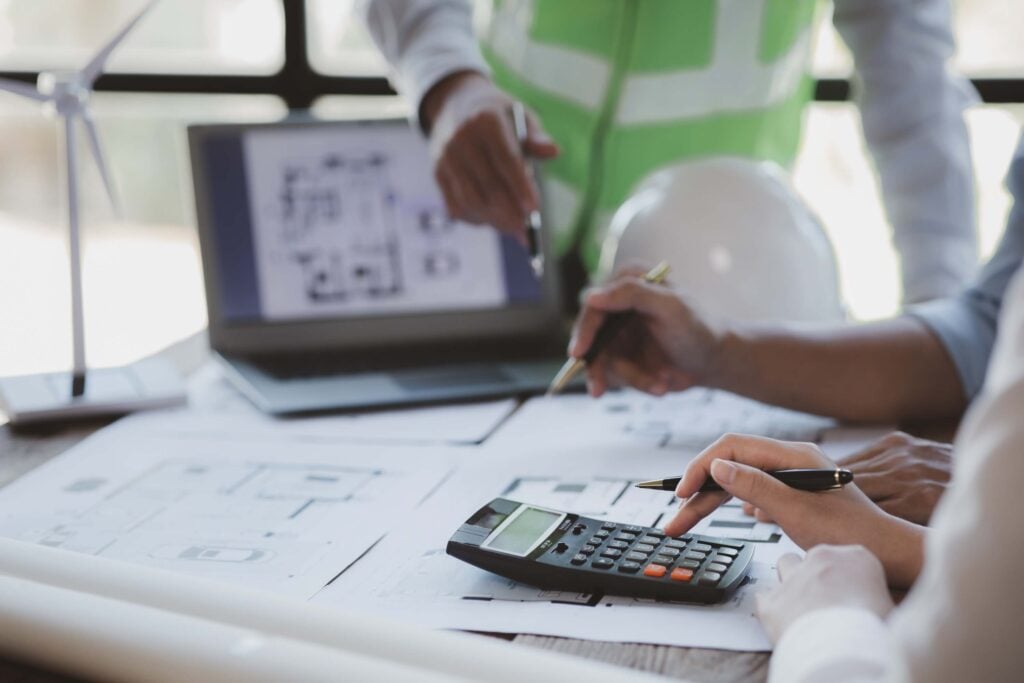 Two people review architectural blueprints on a desk—one uses a calculator while another points at documents, likely discussing a construction project. A laptop, white safety helmet, and small wind turbine model are visible in the background.