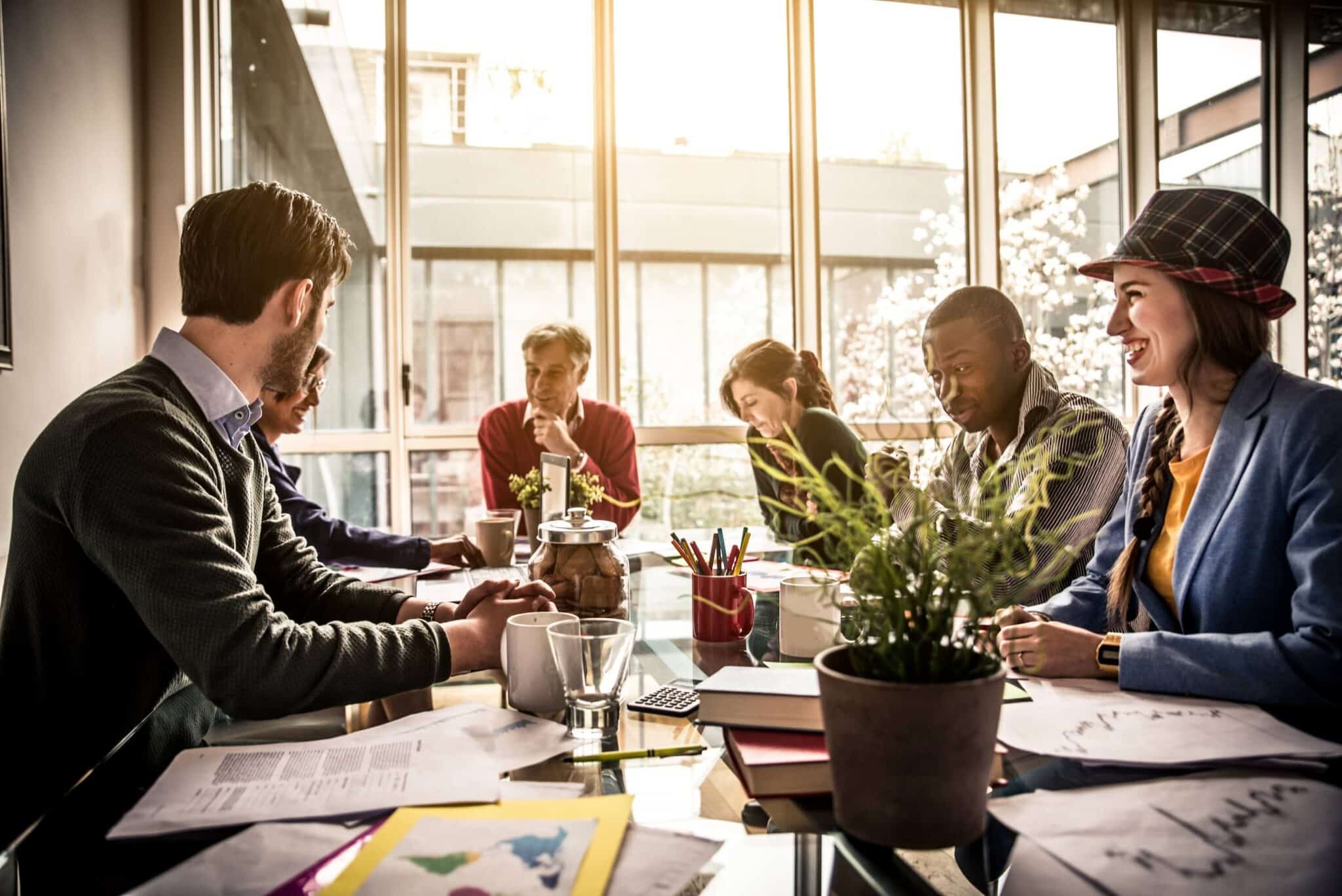 A group of six people sit around a table in a bright ASU 2018 08 office, engaged in discussion. Papers, notebooks, cups, and plants are on the table. Sunlight streams through large windows in the background.