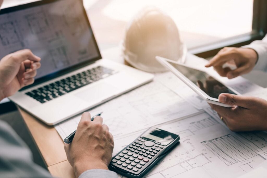 Two people work at a desk on a construction project, reviewing blueprints with a calculator, laptop, and tablet. One uses the tablet while the other takes notes. A hard hat and documents for a construction contract are visible in the background.