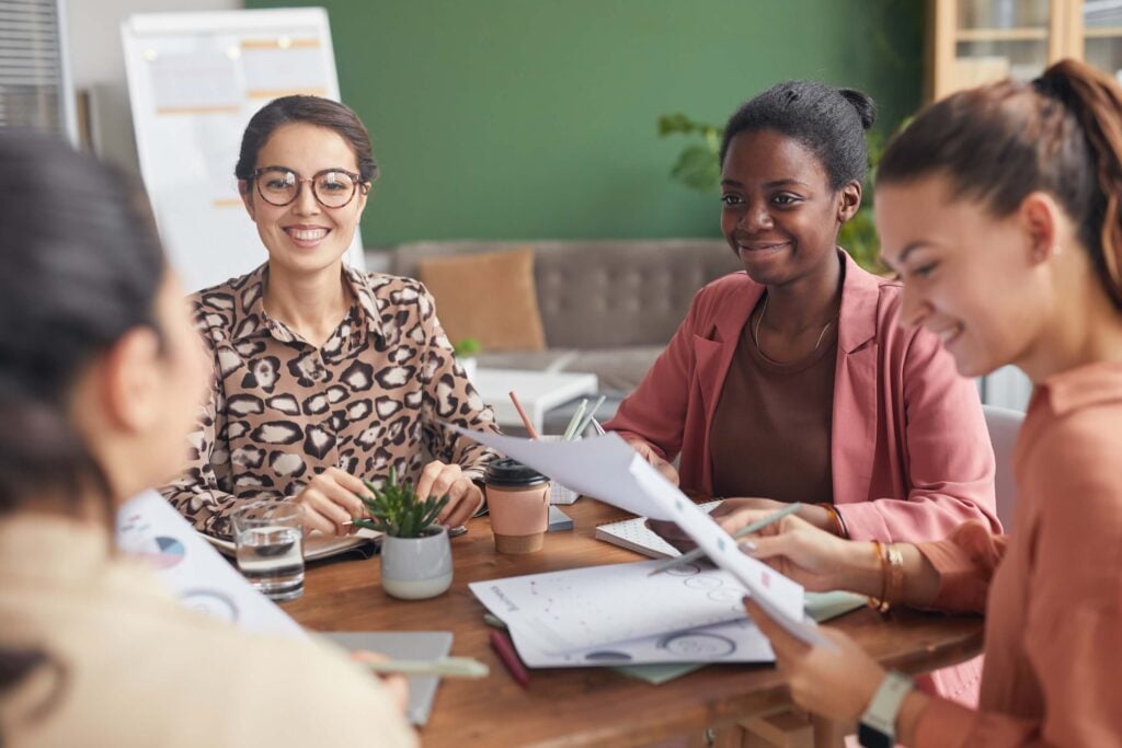 Four women sit around a table in a modern office, smiling and holding papers during a meeting on financial management. Notebooks, coffee cups, and a small plant are on the table. A whiteboard and green wall appear in the background.