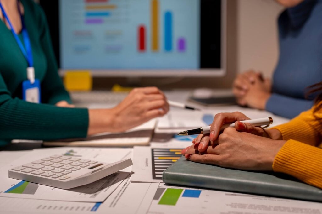 Three people sit at a desk with charts, documents, and a calculator, discussing work and reviewing accounting guidance; a computer screen displaying colorful bar graphs is visible in the background.