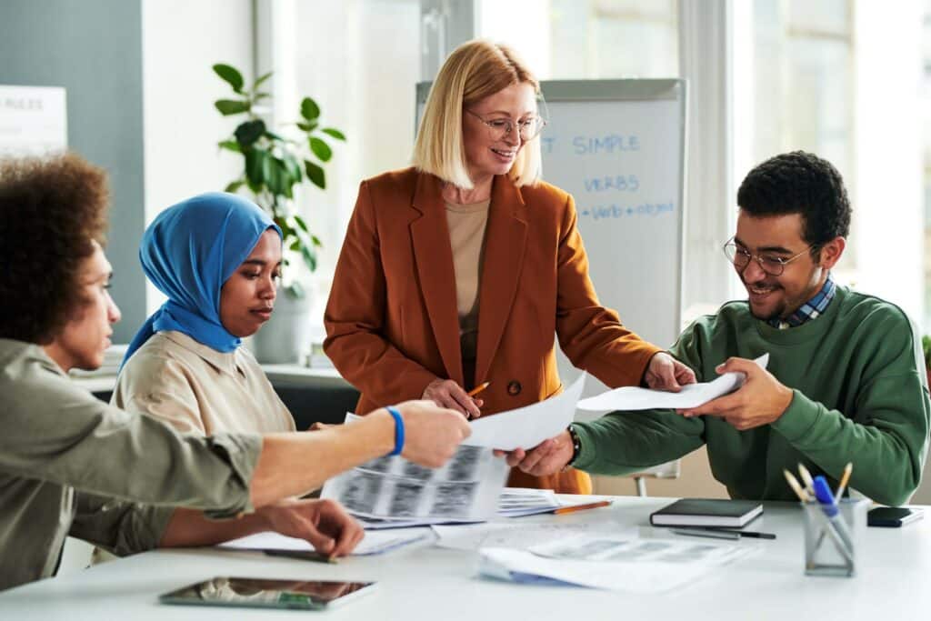 A diverse group of four people sit around a table in an office, exchanging papers and discussing work. One person stands, smiling and facilitating the discussion about new accounting guidance from the Financial Accounting Standards Board.