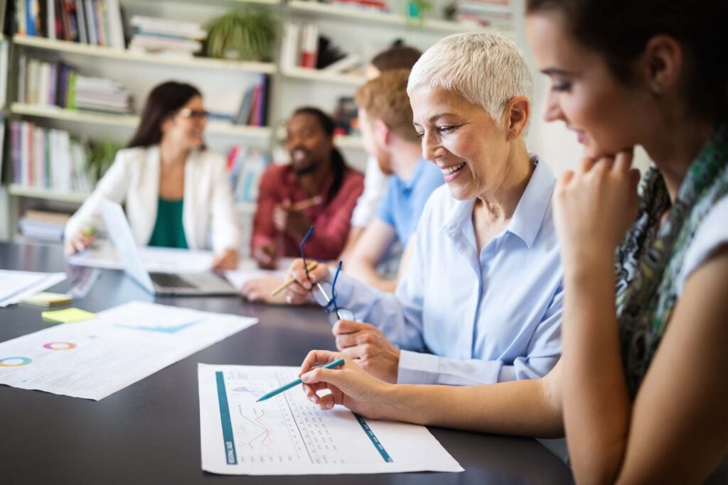 A group of professionals sit at a table in an office discussing documents and financial management charts. Two people in the foreground smile while reviewing a printed graph, with others collaborating near a laptop in the background.
