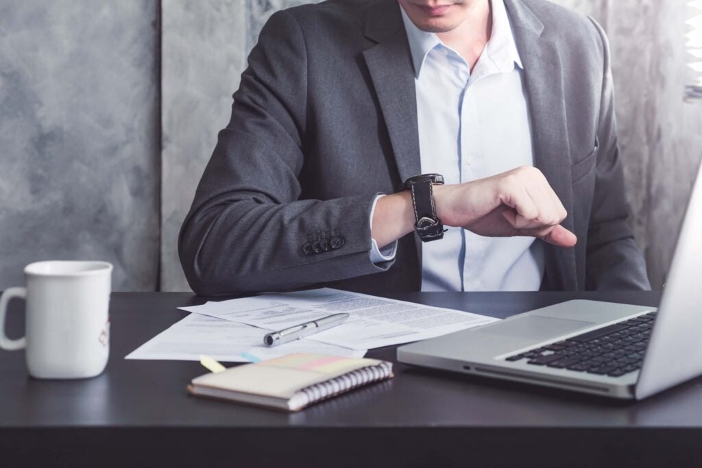 A man in a suit sits at a desk with papers, a laptop, a notebook, and a coffee mug, checking his wristwatch—perhaps comparing time tracking tools like Replicon vs Harvest for their project management features.
