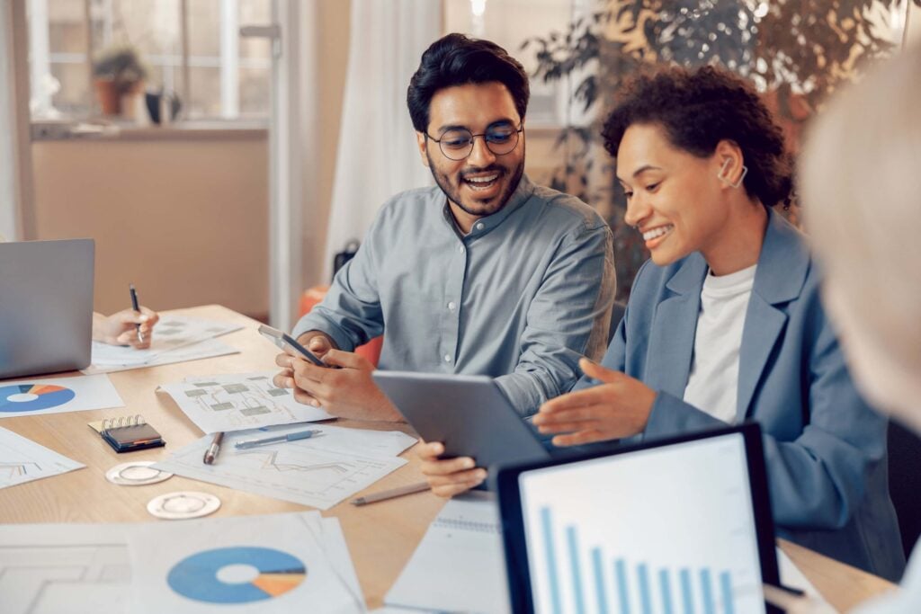 Two colleagues sit at a desk with charts, graphs, and laptops, smiling as they discuss financial management data on a tablet in a bright office setting.