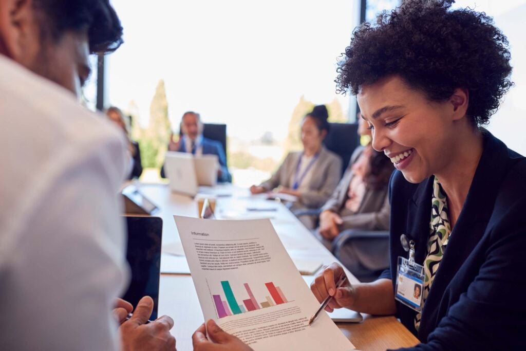 A woman smiles while holding and discussing a paper with colorful bar graphs during a business meeting, possibly explaining recent accounting guidance. Several people are seated around a table, blurred in the background.