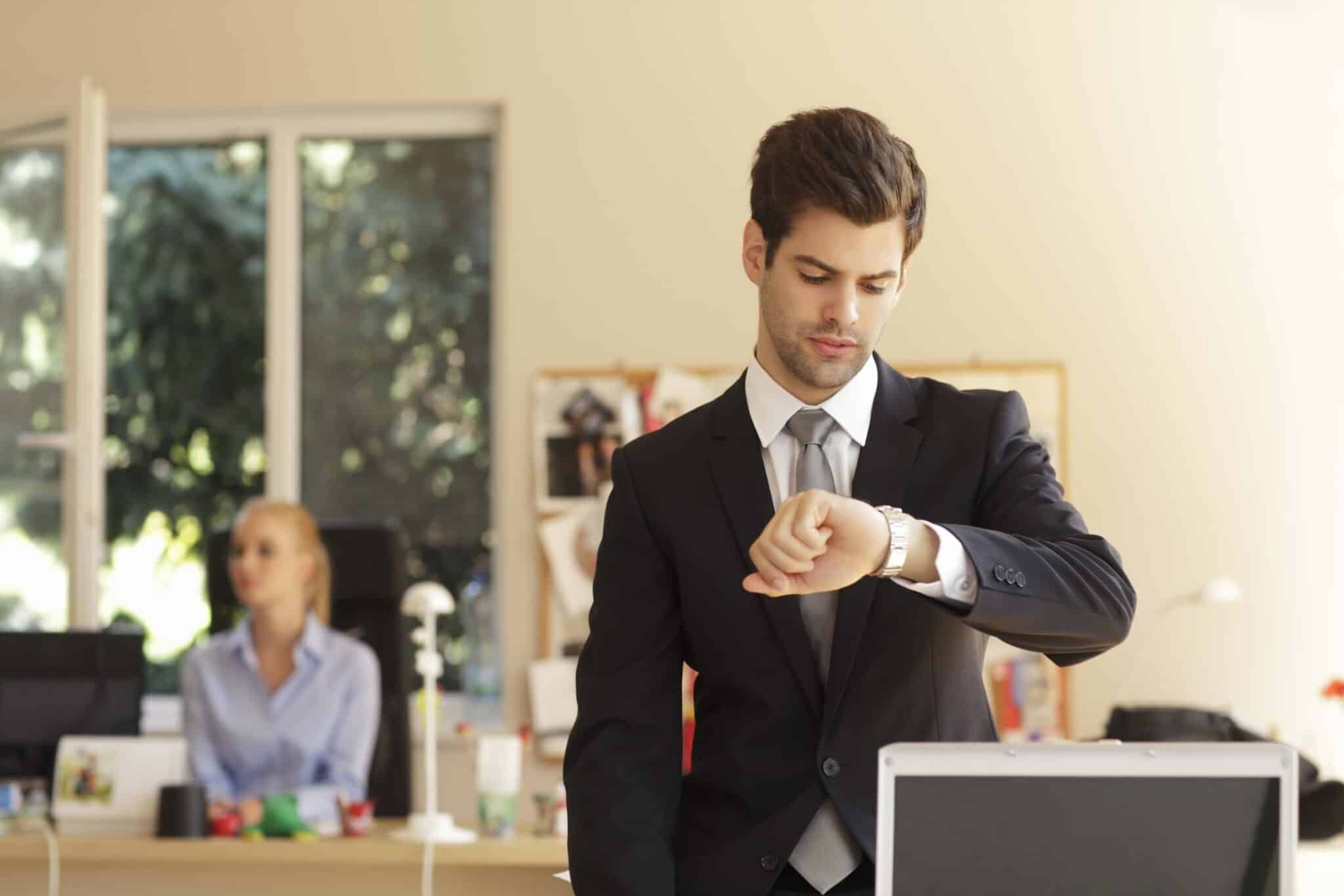 A man in a suit stands in an office, checking his watch—perhaps timing a replicon vs harvest project. In the background, a woman works at her computer as sunlight streams through the window, creating a bright atmosphere.