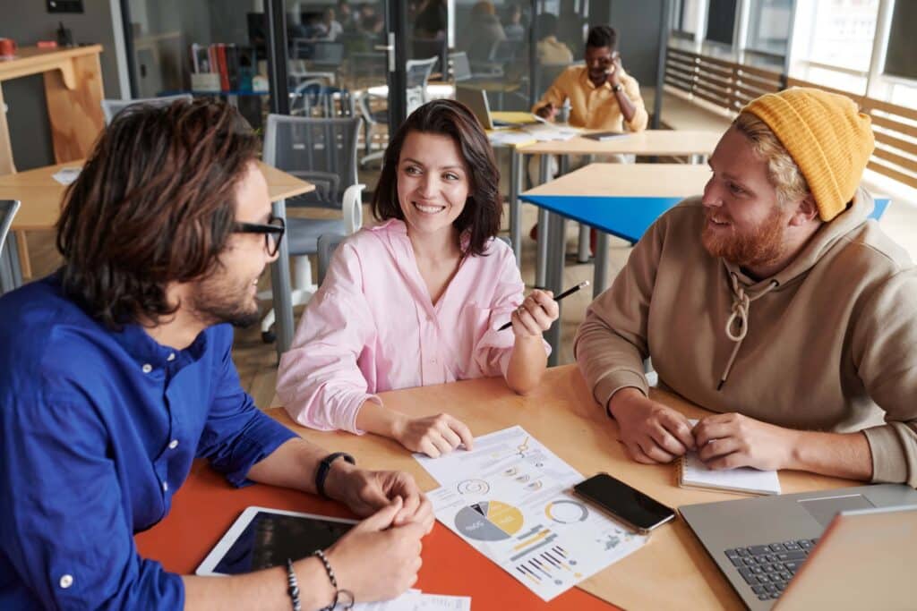 Three young adults sit around a table with charts, a tablet, and a laptop, smiling and discussing Sage Intacct vs Blackbaud. Other people work in the background in a modern, bright office space.