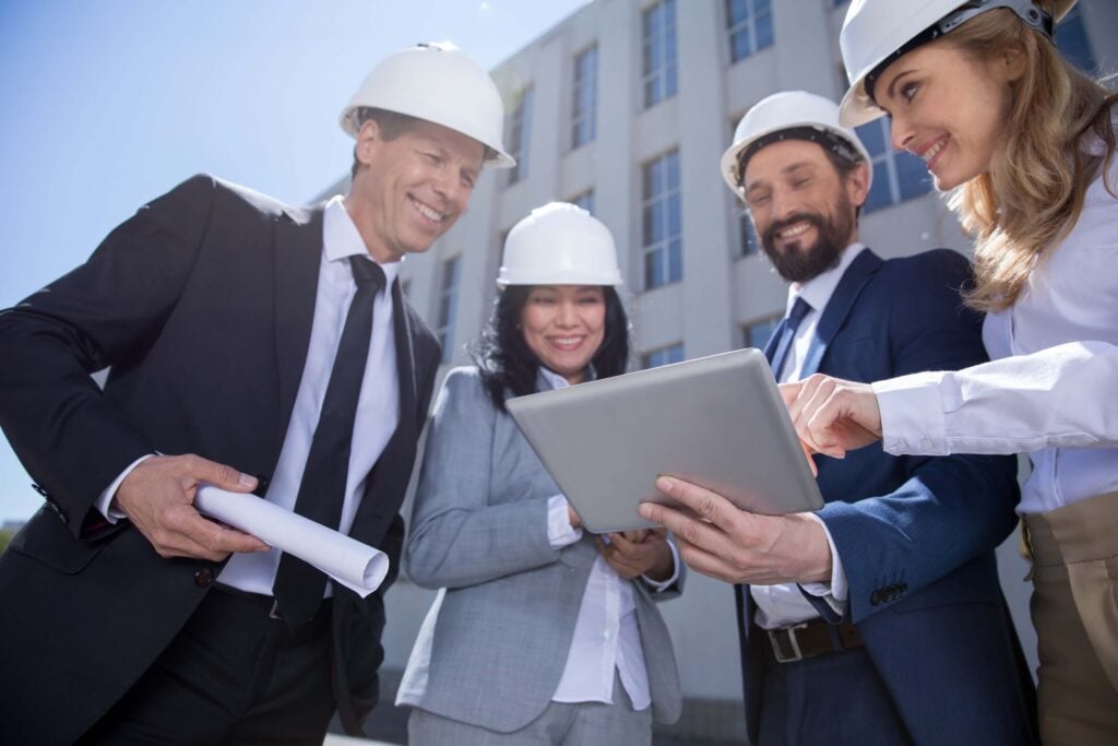 Four professionals in suits and hard hats stand outdoors in front of a building, smiling as they review a construction contract on a tablet held by one woman, while another man holds rolled-up blueprints.