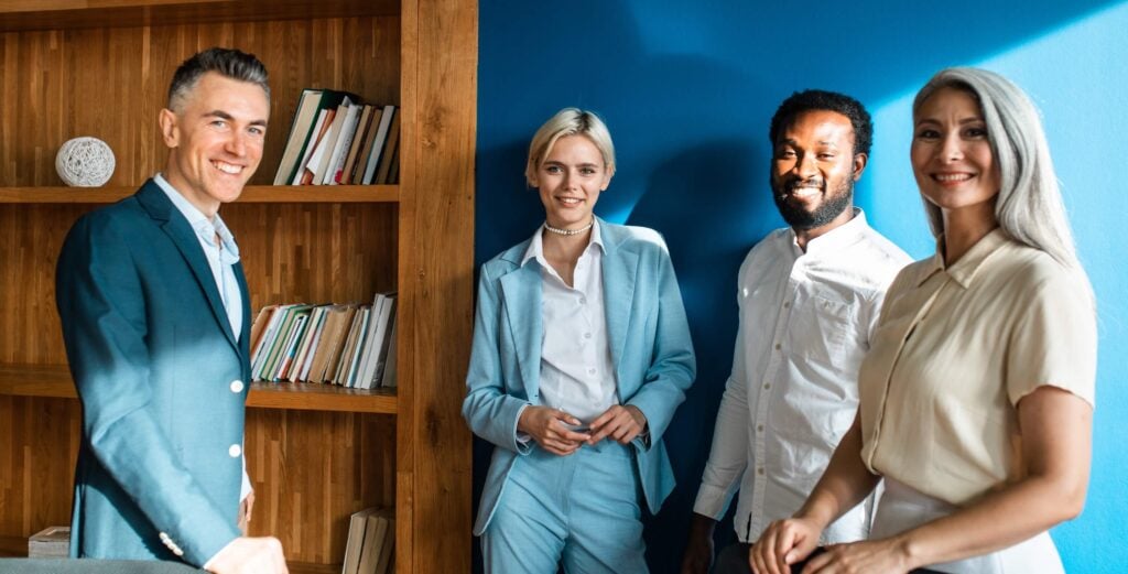 Four professionally dressed people stand smiling in a bright office with blue walls and a wooden bookshelf, discussing project management features to boost efficiency.