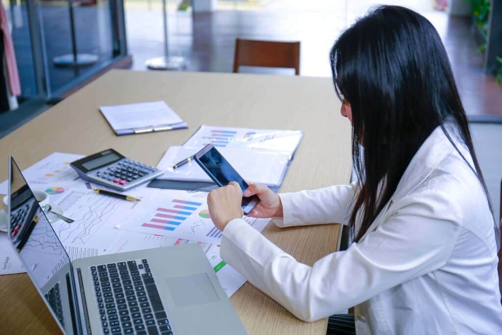 A woman in business attire sits at a desk with a laptop, calculator, and financial documents. She uses her smartphone to snap a photo of charts and graphs—possibly for reporting to the Financial Crimes Enforcement Network on casinos and card clubs.